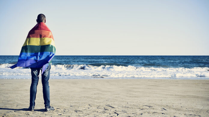a young caucasian man seen from behind wrapped in a rainbow flag